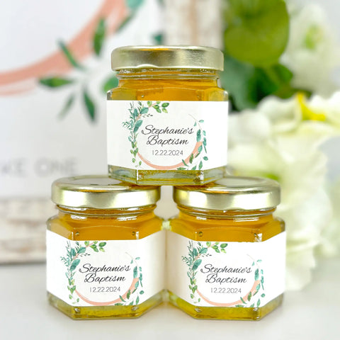 Group of 2 oz honey baptism favors in hexagon jars with gold lid with personalized labels. Labels feature leaf frame. Table decorations in background.