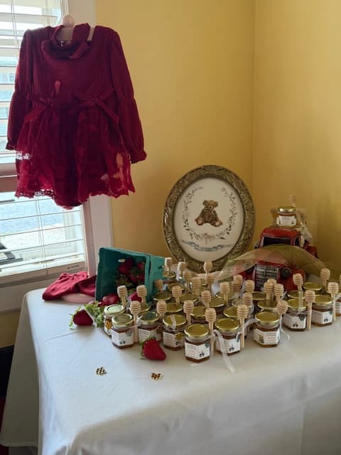 A table covered with small honey jars and topped with a decorative bear-themed picture, set in a room with yellow walls. A dark red dress hangs on a hanger near a window.