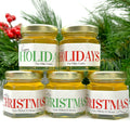 Close up of 2 oz honey holiday Christmas party favor gift jars with gold lid and personalized labels. Labels feature red or green holiday greeting titles, including Merry Christmas or Happy Holidays. Pine tree limbs in background.