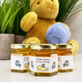 Close up of honey baby shower party favor jars topped with gold lids. Jars have labels featuring classic Winnie the Pooh with his hunny pots and a title that says A Little Yummy For Your Tummy. Table decor and Winnie the Pooh plush toy in background.