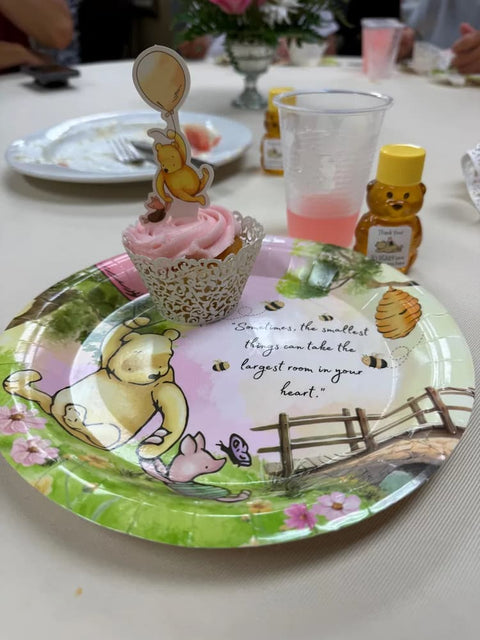 Table place setting with Winnie the Pooh plates, a cupcake, drink, and honey favor bear with yellow cap and Winnie the Pooh label.