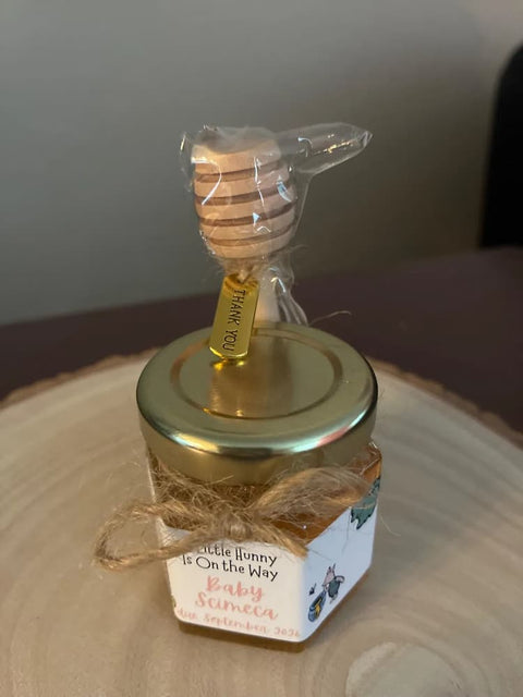 A small labelled glass jar filled with honey, topped with a wooden honey dipper wrapped in clear plastic, and tied with twine on a wooden slice background.