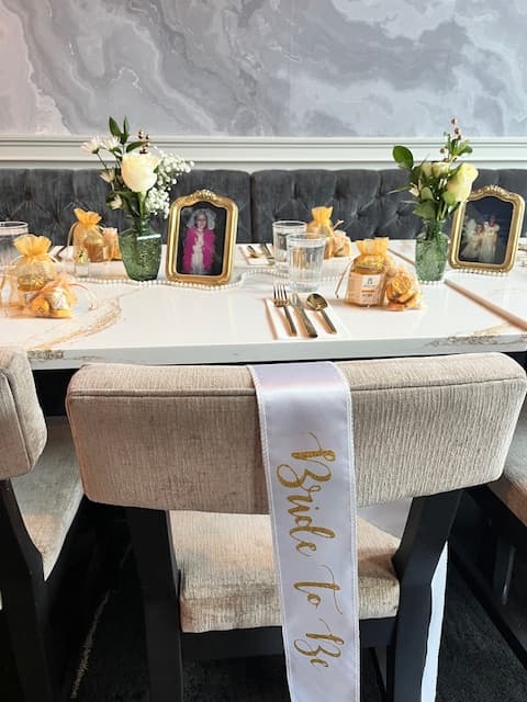 Decorated table with 'Bride to Be' sash on chair, floral arrangements, and framed photos.