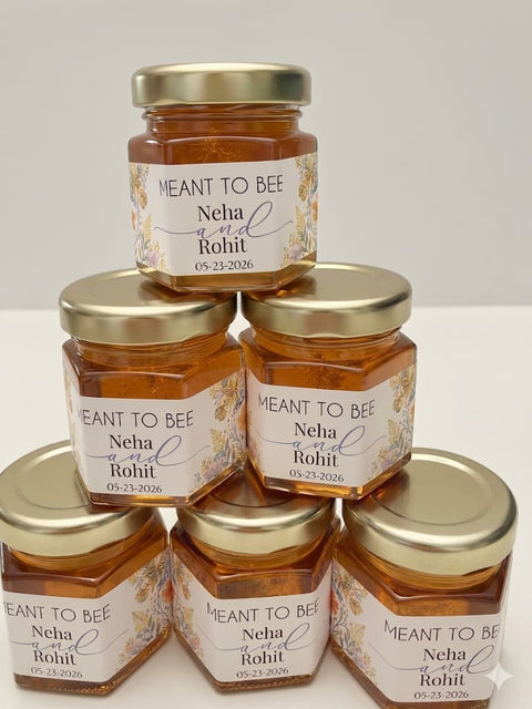 Six small glass jars of honey arranged in a pyramid on a white surface against a light gray background. Each jar has a gold metal lid and a white label with floral accents and the text 'MEANT TO BEE Neha and Rohit' with a date.