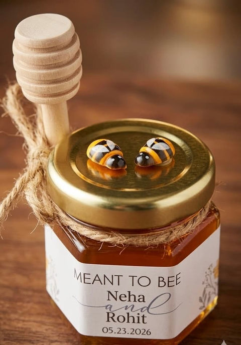 A small glass jar of honey with a gold lid, twine, and wooden honey dipper, featuring two bee-shaped decorations on the lid. The hexagonal jar has a white label reading 'MEANT TO BEE Neha and Rohit.