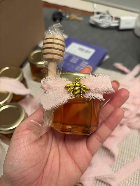 Jar of honey with bee charm, ribbon, and honey dipper all held by a hand.