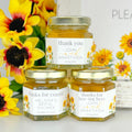 Close up of 2 oz honey thank you wedding party shower favors on hexagon jars with gold lid with personalized labels. Labels feature sunflower design. Label titles shown are Thank You, Thanks for Bee-ing Here, and Thanks for Coming. Table decorations in background.