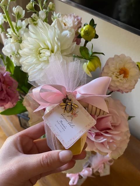 Small honey jar with a bee charm held by a hand, surrounded by flowers.