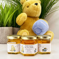 Close up of honey baby shower party favor jars topped with gold lids. Jars have labels featuring classic Winnie the Pooh with his hunny pots and a title that says A Little Yummy For Your Tummy. Table decor and Winnie the Pooh plush toy in background.