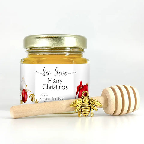 2 oz hexagon honey favor. Jar is filled with honey and has gold lid, 3" wooden honey dipper, gold bee charm, and personalized Christmas ornament label with title Bee-lieve.