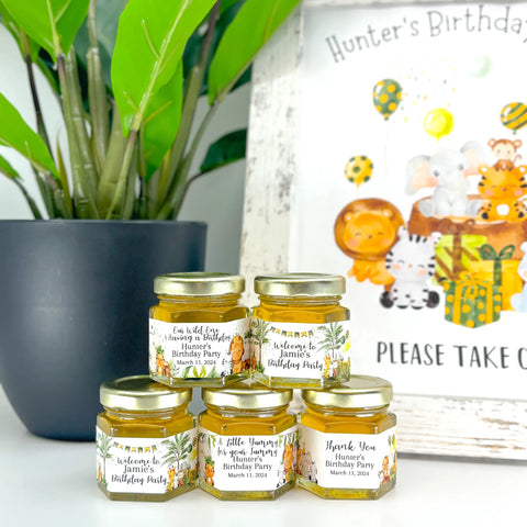 Party favor table decor is shown including a plant and honey birthday party favor jars topped with gold lids. Jars have labels featuring jungle animals with party decorations and a title. Titles include Our Wild One is Having a BIrthday, Welcome to Jamie's Birthday Party, A Little Yummy for your Tummy, and Thank You. Matching free printable sign is also shown.