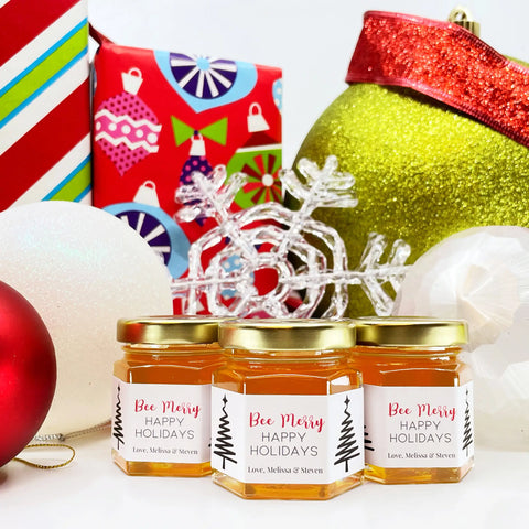 Close up of 2 oz honey holiday Christmas party favor gift jars with gold lid and personalized labels. Labels feature one line Christmas trees and Bee Merry title. Christmas presents and ornaments in background.