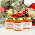 Close up of 2 oz honey holiday Christmas party favor gift jars with gold lid and personalized labels. Labels feature Christmas ornament design and title saying wishing you a sweet holiday. Pine tree and ornaments in background.