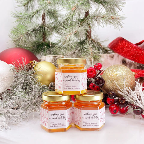 2 oz honey holiday Christmas party favor gift jars shown with gold lid and personalized Christmas ornament labels. Labels feature title saying wishing you a sweet holiday.. Christmas ornaments and tree in background.