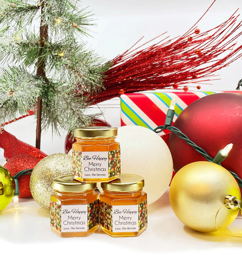 2 oz honey holiday Christmas party favor gift jars shown with gold lid and personalized holiday lights design on labels. Labels feature Bee Happy title. Christmas ornaments, lights, tree, and presents in background.