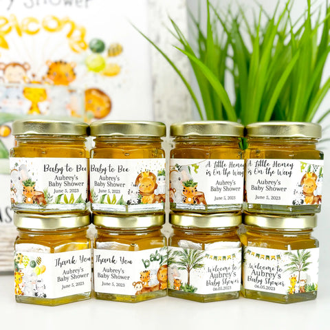 Close up of honey baby shower favor jars topped with gold lids. Jars have labels featuring jungle animal designs and a variety of titles. Titles shown include Baby to Bee, A Little Honey is On the Way, Thank You, and a Welcome title. Table decor in background.