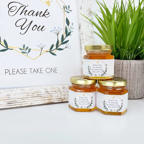 2 oz honey thank you wedding party shower favors on hexagon jars with gold lid and labels. Labels feature geometric design with title that says Thank You for sharing our bee-utiful day with us. Free matching printable table sign in background.