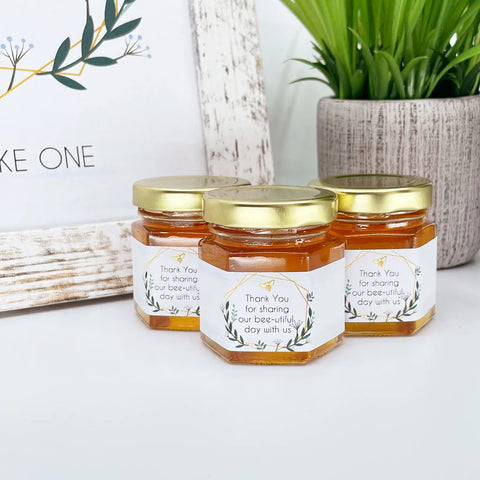 Close up of 2 oz honey thank you wedding party shower favors on hexagon jars with gold lid and labels. Labels feature geometric frame design and title that says Thank You for sharing our bee-utiful day with us. Table decorations in background.