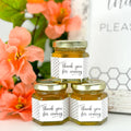 Close up of 2 oz honey thank you wedding party shower favors on hexagon jars with gold lid and labels. Labels feature black and white honeycomb design and Thank You for Coming title. Table decorations in background.