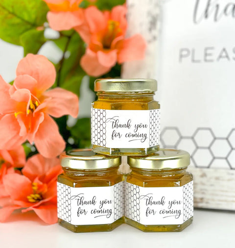 Close up of 2 oz honey thank you wedding party shower favors on hexagon jars with gold lid and labels. Labels feature black and white honeycomb design and Thank You for Coming title. Table decorations in background.