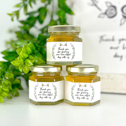 Close up of 2 oz honey nee-utiful date wedding party shower favors on hexagon jars with gold lid and labels. Labels feature rose laurel design and title stating Thank you for sharing our bee-utiful day with us. Table decorations in background.