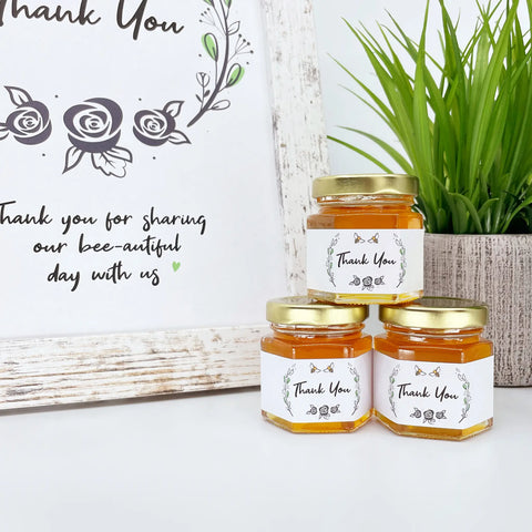 2 oz honey thank you wedding party shower favors on hexagon jars with gold lid and labels. Labels feature rose laurel design with Thank You title. Free matching printable table sign in background.