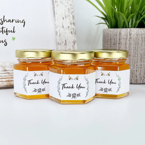 Close up of 2 oz honey thank you wedding party shower favors on hexagon jars with gold lid and labels. Labels feature rose laurel design and Thank You title. Table decorations in background.