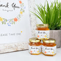 2 oz honey thank you wedding party shower favors on hexagon jars with gold lid and labels. Labels feature wildflower design with Thank You title. Free matching printable table sign in background.