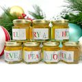 Side view of 2 oz honey holiday Christmas party favor gift jars with gold lid and personalized labels. Labels feature multicolored holiday greeting titles. Images shown on sides include silver snowflakes, blue snowflakes, and various Christmas trees. Christmas decorations in background.