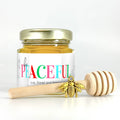 2 oz hexagon honey favor. Jar is filled with honey and has gold lid, 3" wooden honey dipper, gold bee charm, and personalized multicolored holiday greeting design on label with title Bee Peaceful.