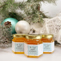 Close up of 2 oz honey holiday Christmas party favor gift jars with gold lid and personalized labels. Labels feature snowflake design and Bee Joyful title. Pine tree and ornaments in background.