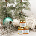 2 oz honey holiday Christmas party favor gift jars shown with gold lid and personalized snowflake labels. Labels feature Bee Joyful title. Christmas ornaments and pine tree in background.