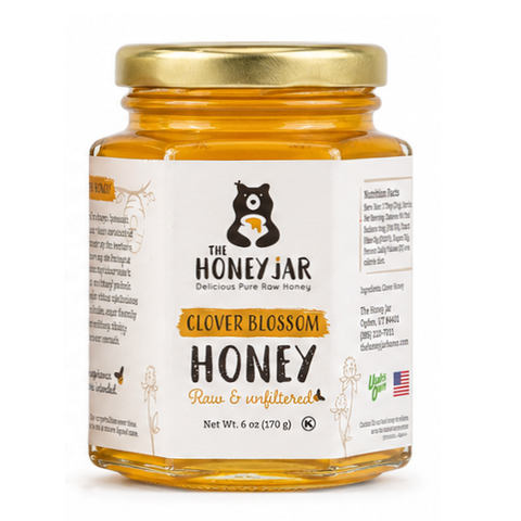 A jar of Clover Blossom Honey from The Honey Jar, featuring a white label with black and gold text, a gold metal lid, and the honey visible through the clear glass jar against a white background.
