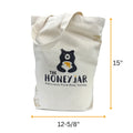 A cream colored tote bag with a bear and a honey pot. The Honey Jar Delicious Pure Raw Honey printed on it. Dimensions shown are 15" high and 12-5/8" wide.