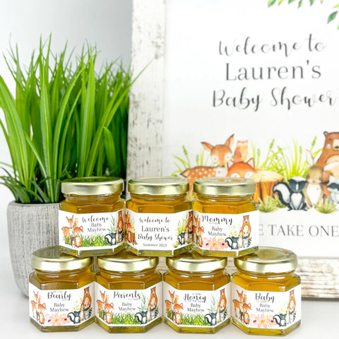 Baby Shower favor table decor shown with 2 oz hexagon favor jars filled with honey and topped with gold lid. Jars have labels featuring a variety of woodland animal designs. Label titles include Baby to Bee, A Little Honey is on the Way, Parents to Bee, We Can Bearly Wait, Mommy to Bee, and two Welcome labels. Table shows free matching printable sign and plant in background.