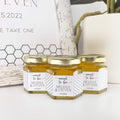 Close up of honey wedding favors with personalized honeycomb labels, 2 oz hexagon jars, and gold lids. Title on label is Meant to Bee. Table decorations in background