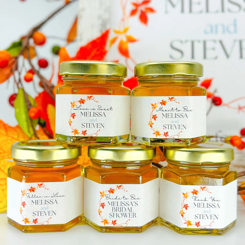 2 oz honey wedding favors in hexagon jars with gold lids and personalized labels. Labels feature a fall leaf frame design and various titles. Titles shown include Love is Sweet, Meant to Bee, Fallen in Love, Bride to Bee, and Thank You.