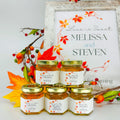 Honey favor table display featuring 2 oz hexagon jars filled with honey and topped with gold lids. Labels on jars show fall leaf frame design and personalized titles, including Love is Sweet, Fallen in Love, Bride to Bee, Thank You, and Meant to Bee. Free matching printable table sign with Love is Sweet title is shown behind jars.