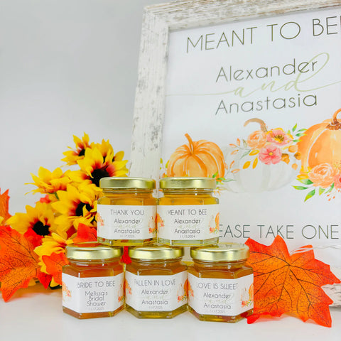 Example of honey favor table decor with 2 oz honey favors in the forefront. Jars are topped with gold lids and have autumn pumpkin themed labels with varying titles. Titles include Thank You, Meant to Bee, Bride to Bee, Fallen in Love, and Love is Sweet. A free matching printable table sign with the title Meant to Bee is in the background.