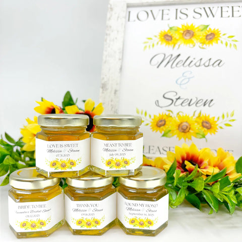 Honey wedding favor table setting with 2 oz hexagon jars filled with honey, topped with gold lids, and showing personalized sunflower bunch labels. Titles shown on labels are Love is Sweet, Meant to Bee, Bride to Bee, Thank You, and Found My Honey. Table decor includes free matching printable sign with title Love is Sweet and floral arrangement.