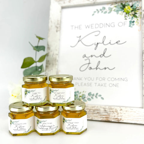 Gold Eucalyptus themed wedding favor table with 2 oz honey wedding favors in front. Jars are topped with gold lids and have personalized gold eucalyptus theme labels with titles: Thank You, Bee Our Guest, Spread the Love, Bride to Bee, and Found My Honey. Free matching printable sign is in the background.