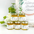 close up of leaf frame honey wedding favors with personalized labels. Titles include spread the love, thank you, found my honey, bride to bee, bee our guest, and more. Flowers and table decorations in background.