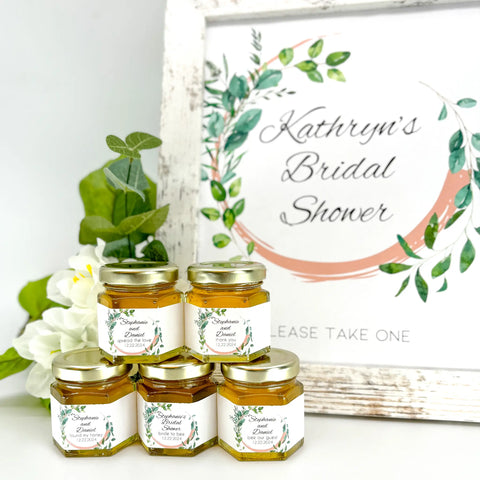 wedding favor table setting including free printable sign and stack of honey wedding favors with personalized leaf frame label. Possible titles include thank you, spreaad the love, found my honey, bride to bee, bee our guest, and more. Jars are filled with clover honey.