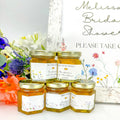 close up of meadow wedding favors with personalized labels, honey, and gold lids. Title options include Bee Our Guest, Found My Honey, Bride to Bee, Welcome, Love is Sweet, and more. Showcasing different flower color options: multi, yellow, blue, red/pink, and purple.