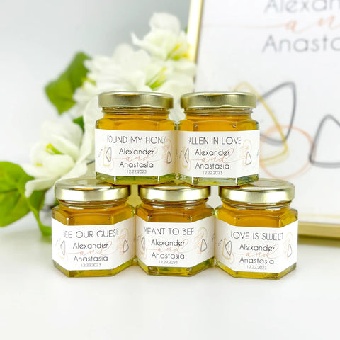 Close up of 2 oz wedding favors: hexagon jars, gold lids, and personalized labels with rose gold geometric theme. Label titles include Found My Honey, Fallen in Love, Bee Our Guest, Meant to Bee, Love is Sweet, and more.