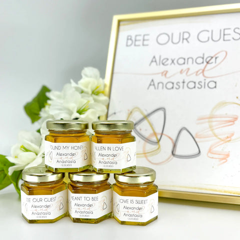 Rose gold geometric 2 oz honey wedding favors with gold lids and personalized labels. Label titles include Found My Honey, Fallen in Love, Bee Our Guest, Meant to Bee, and Love is Sweet. Matching printable sign with Bee Our Guest title is in the background.